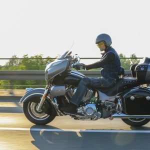 A close-up of a man wearing a helmet and safety gear while riding a motorcycle on the highway. He is near a guardrail.