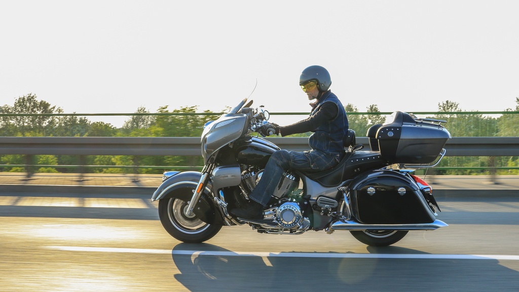 A close-up of a man wearing a helmet and safety gear while riding a motorcycle on the highway. He is near a guardrail.
