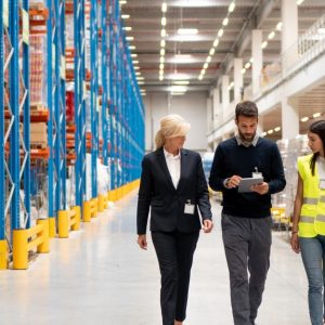 Two managers in business casual clothing in a warehouse with a worker. They are walking next to industrial shelving.