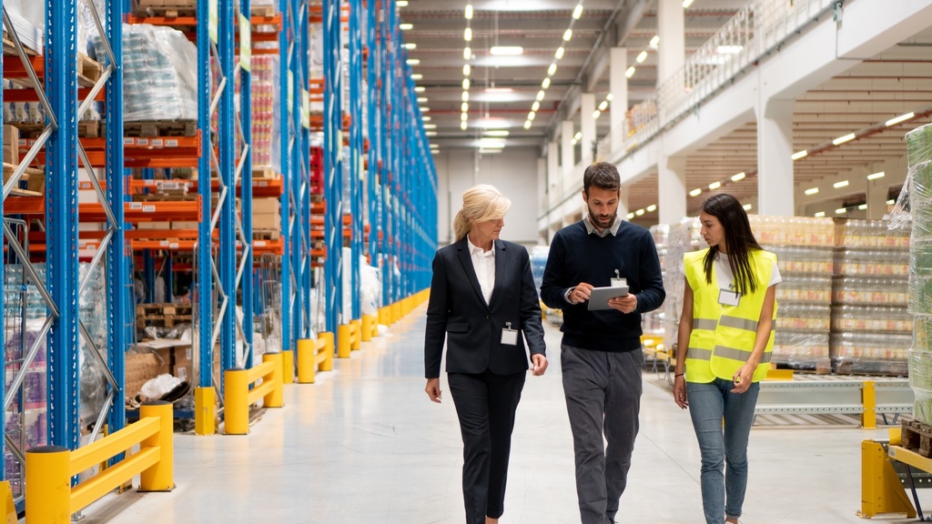 Two managers in business casual clothing in a warehouse with a worker. They are walking next to industrial shelving.