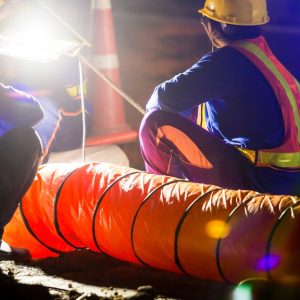 A close-up of two construction workers wearing hard hats and safety vests while working under a large light at night.