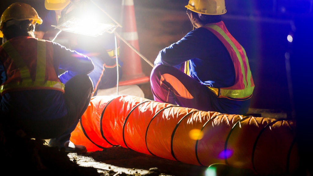 A close-up of two construction workers wearing hard hats and safety vests while working under a large light at night.