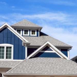 A multi-story blue beach home with a multi-tiered shingle roof with white-framed windows against a blue sky.