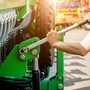 A close-up of the hands of a man holding a long screwdriver to tighten the bolts on the side of a large harvester.