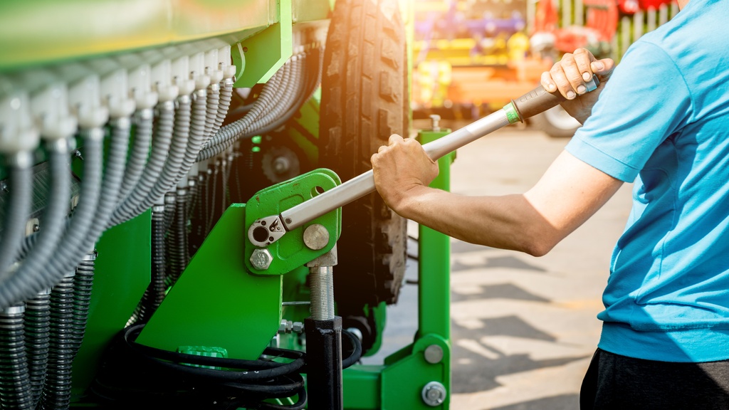 A close-up of the hands of a man holding a long screwdriver to tighten the bolts on the side of a large harvester.
