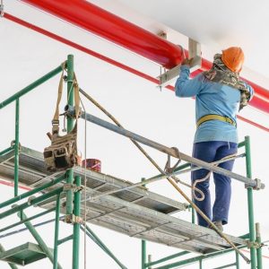A person wearing an orange hard hat and blue shirt stands on scaffolding to paint a steel pipe red.