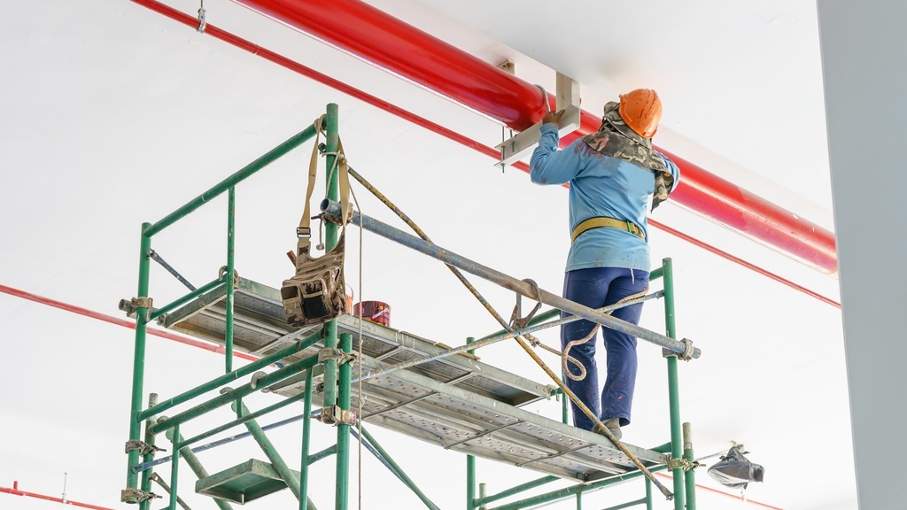 A person wearing an orange hard hat and blue shirt stands on scaffolding to paint a steel pipe red.