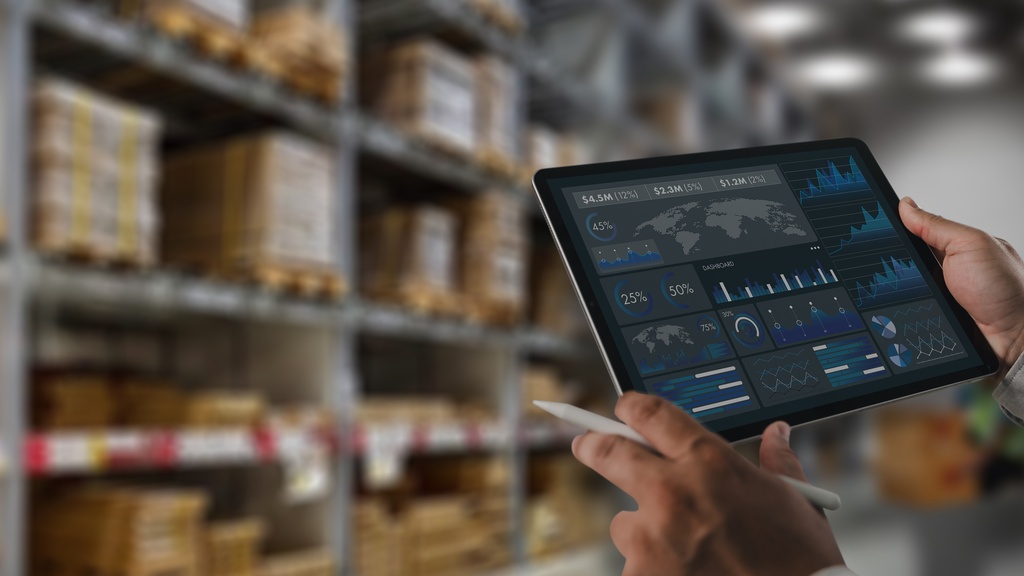 A person is standing in a warehouse while they’re holding a tablet in their hands. They have a white stylus in their hand as well.