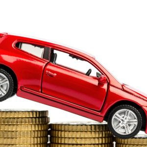 A close-up of a small, red hatchback toy car pointed downward while on stacks of descending gold coins.