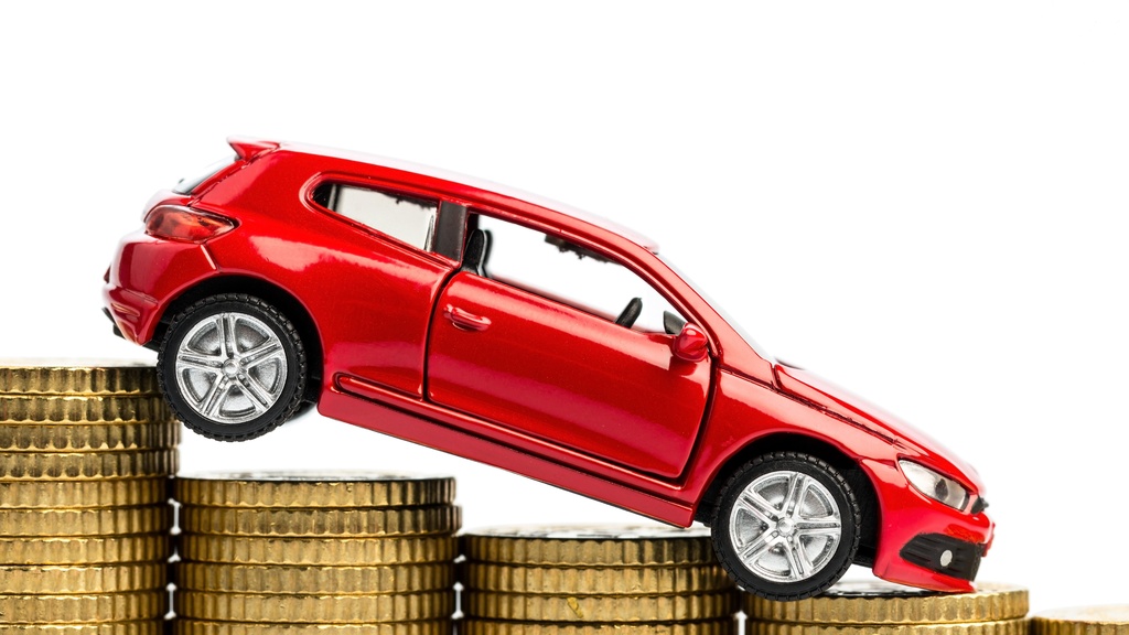 A close-up of a small, red hatchback toy car pointed downward while on stacks of descending gold coins.