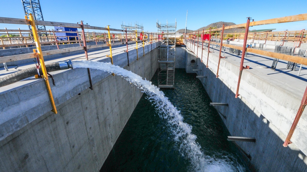 a black hose releasing water into a concrete basin with water, wooden beams, and yellow supports in the back.