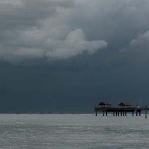 Dark storm clouds loom over a calm Gulf Coast bay as a wooden pier stretches across the water in the distance.
