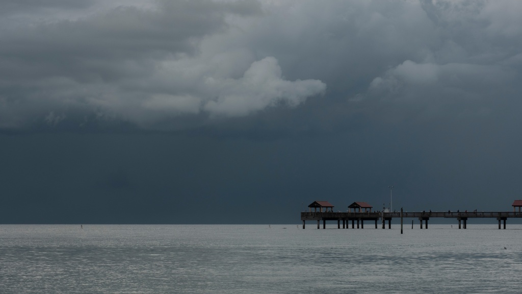 Dark storm clouds loom over a calm Gulf Coast bay as a wooden pier stretches across the water in the distance.