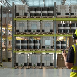 A man wearing a yellow safety vest and a hard hat stands in front of chemical drums and IBC totes stacked on shelving.