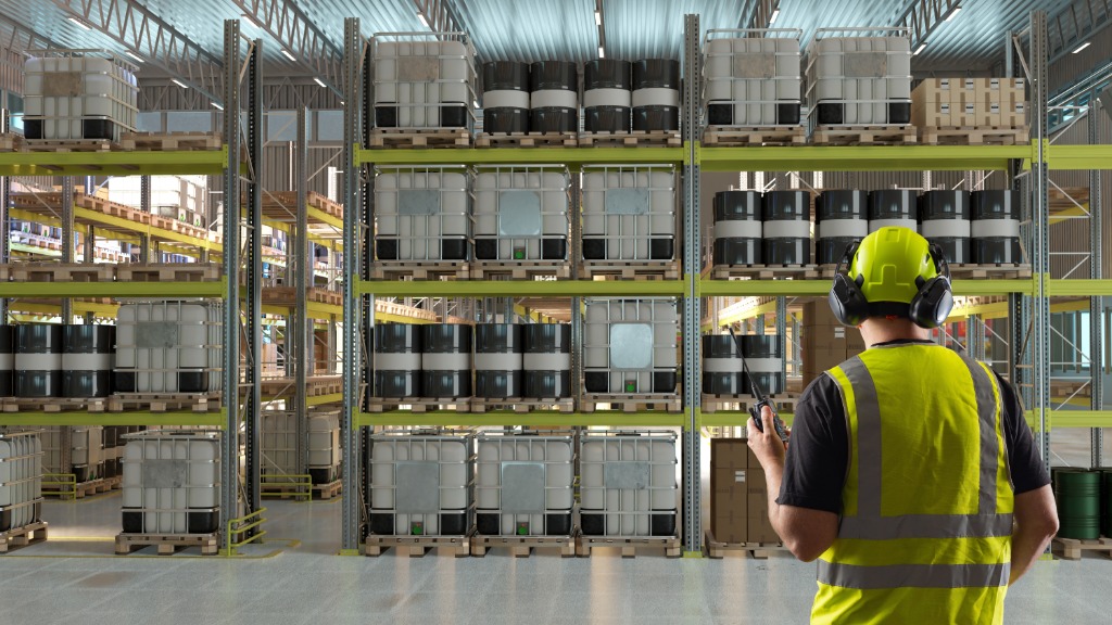 A man wearing a yellow safety vest and a hard hat stands in front of chemical drums and IBC totes stacked on shelving.