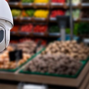 A white dome security camera mounted on a wall, overlooking a blurred grocery store produce section.