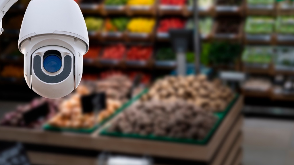 A white dome security camera mounted on a wall, overlooking a blurred grocery store produce section.