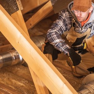 A man wearing gloves kneeling in an attic beside metal ductwork among wooden beams and loose insulation.
