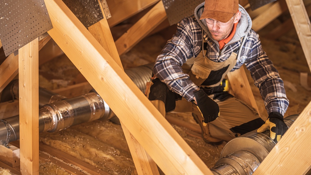 A man wearing gloves kneeling in an attic beside metal ductwork among wooden beams and loose insulation.