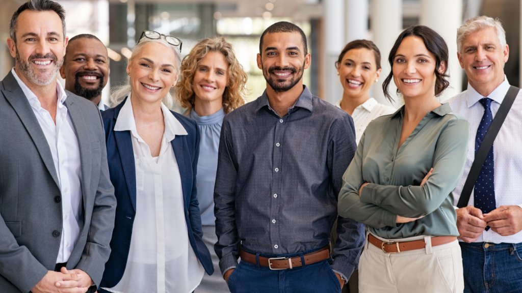 Eight professionally dressed adults of various ages standing and smiling in a brightly lit office space.
