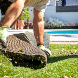 A homeowner wearing gloves rolls up artificial turf beside a backyard pool as grass clippings scatter in the sun.