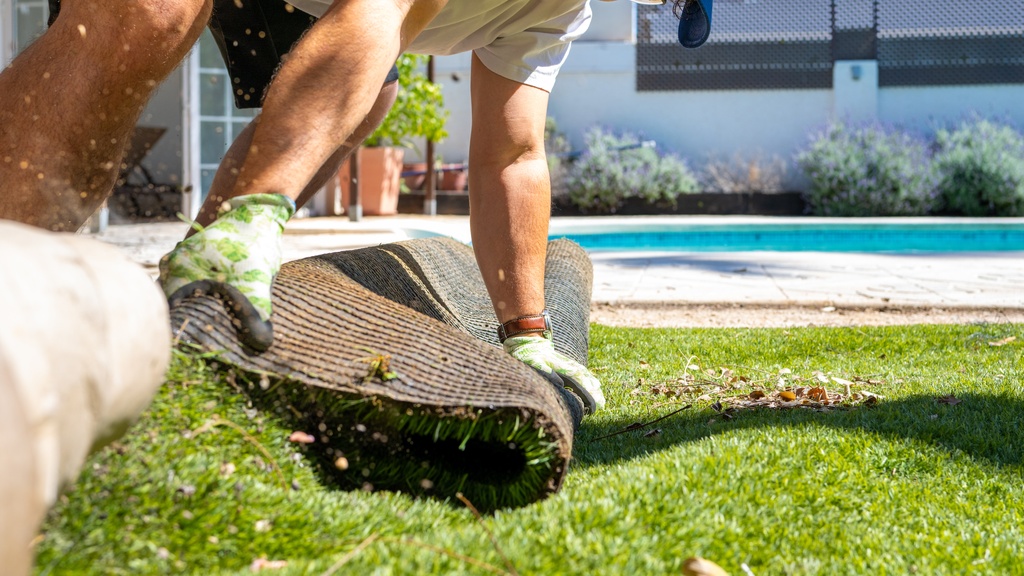 A homeowner wearing gloves rolls up artificial turf beside a backyard pool as grass clippings scatter in the sun.