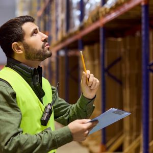 A warehouse employee inspecting shelves full of inventory. The employee holds a clipboard and pencil.