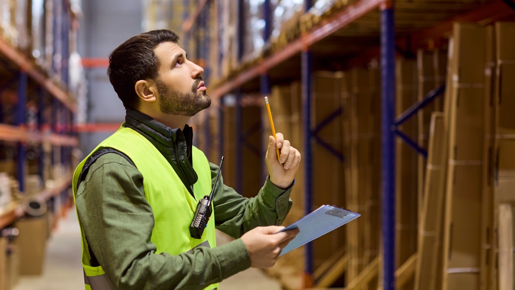 A warehouse employee inspecting shelves full of inventory. The employee holds a clipboard and pencil.