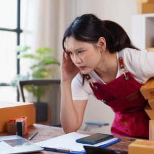 A female employee in a red apron leans over a desk with one hand at her temple, holding stacked boxes near a laptop.