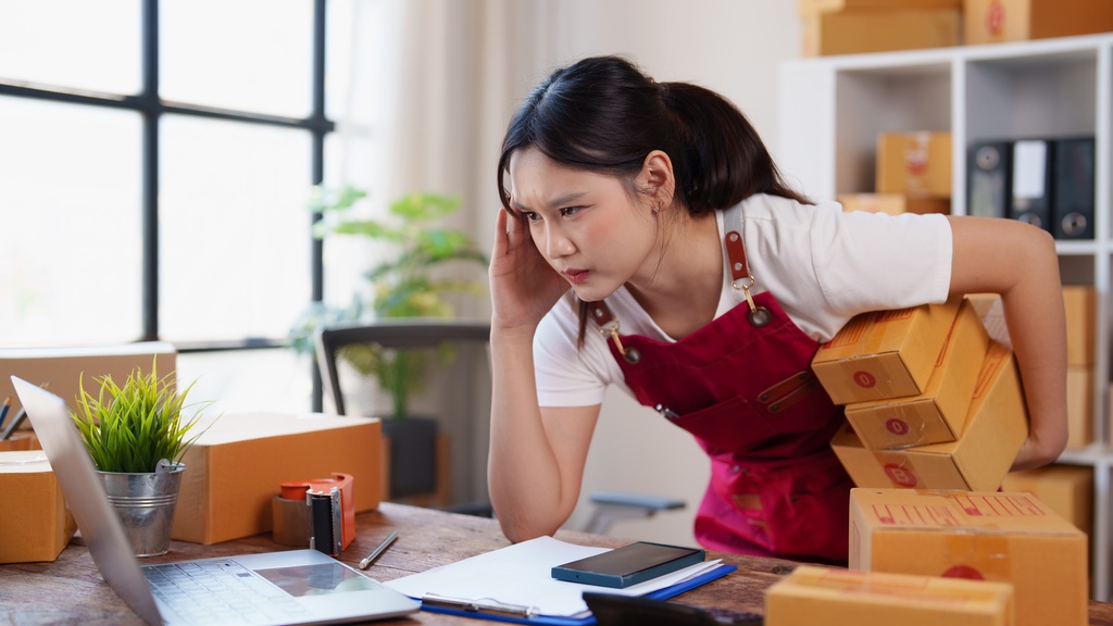 A female employee in a red apron leans over a desk with one hand at her temple, holding stacked boxes near a laptop.