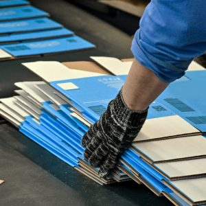 A person holds stacked cardboard boxes inside a production setting with paper materials and containers visible.