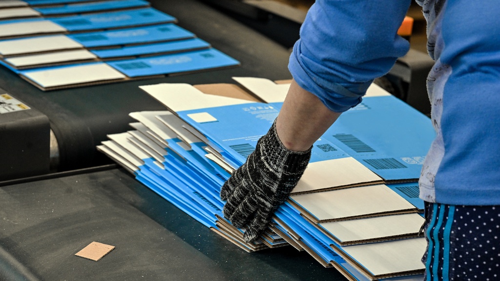 A person holds stacked cardboard boxes inside a production setting with paper materials and containers visible.