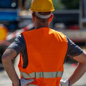 A worker in a yellow hard hat and orange safety vest standing with hands on hips between road barriers and trucks.