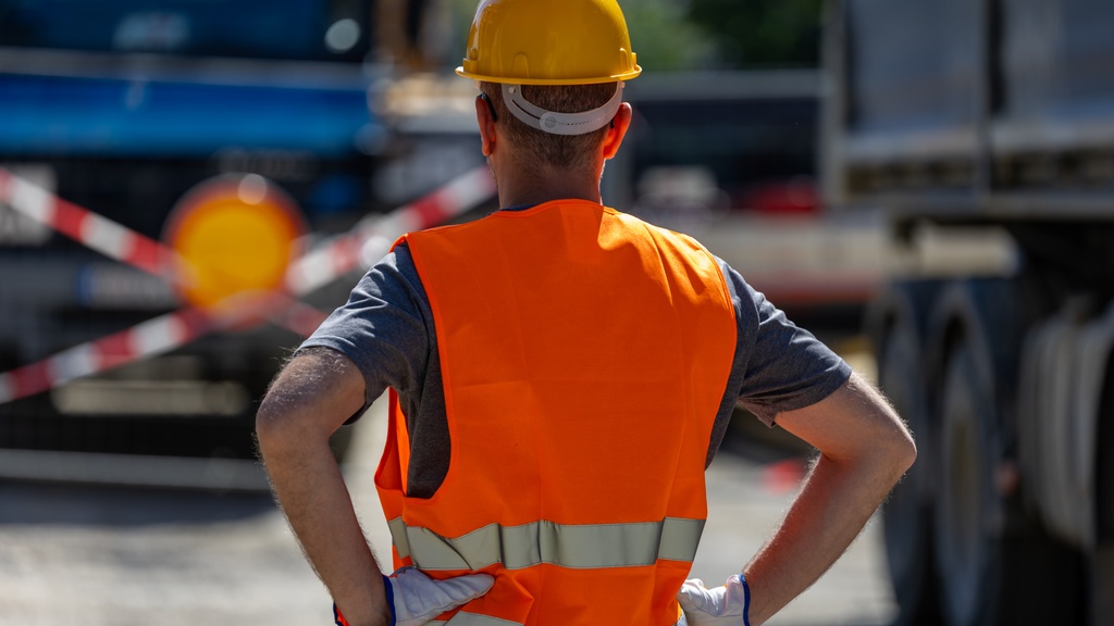 A worker in a yellow hard hat and orange safety vest standing with hands on hips between road barriers and trucks.