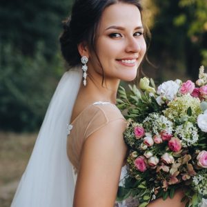 Smiling bride in a white wedding gown and veil holds a lush pink and white floral bouquet in an outdoor garden setting.