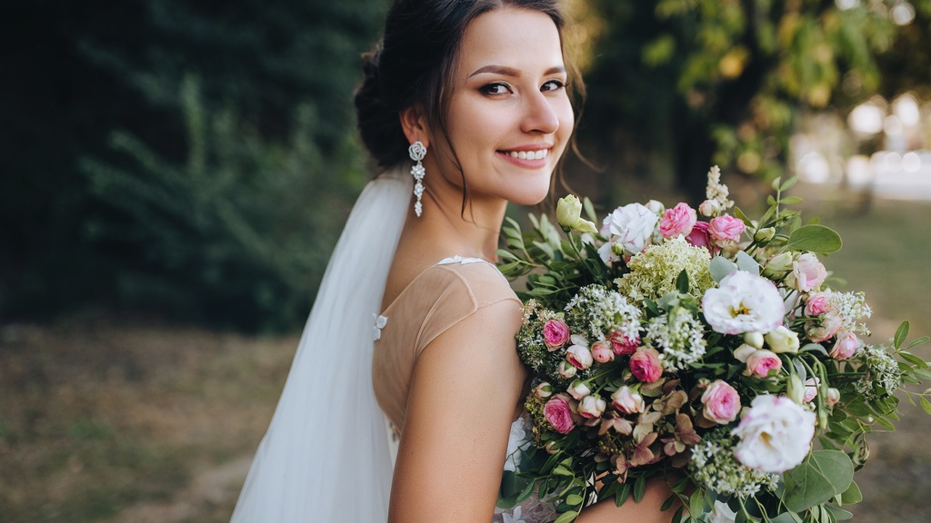 Smiling bride in a white wedding gown and veil holds a lush pink and white floral bouquet in an outdoor garden setting.