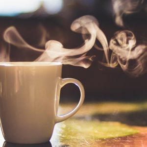 A close-up of a morning cup of coffee with steam flowing from the top sits on a table with the sunrise hitting it.