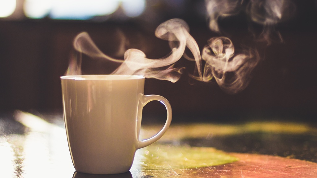 A close-up of a morning cup of coffee with steam flowing from the top sits on a table with the sunrise hitting it.