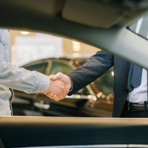 Two people shake hands beside a car indoors, with a steering wheel and front seat visible in the foreground.
