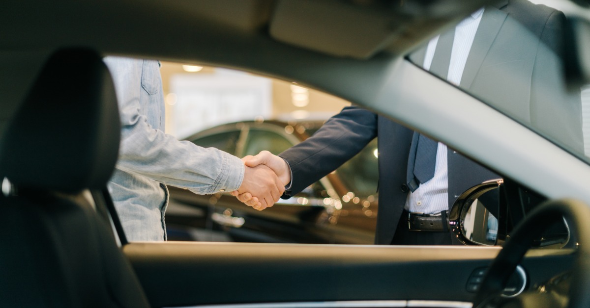 Two people shake hands beside a car indoors, with a steering wheel and front seat visible in the foreground.