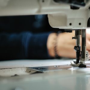 A close-up of the needle of an industrial sewing machine with white thread and two hands in the background.
