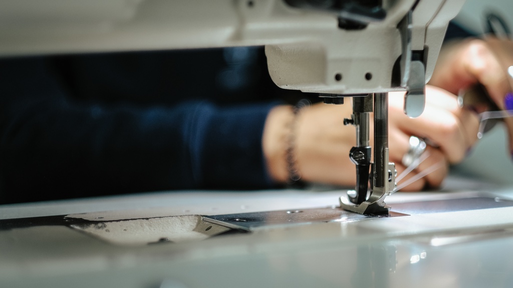 A close-up of the needle of an industrial sewing machine with white thread and two hands in the background.