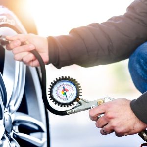 A close-up of a man in jeans and a brown jacket crouching to use a tire pressure gauge on the valve stem of a car tire.