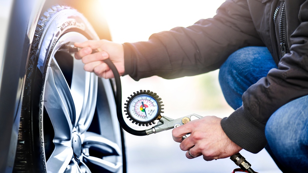 A close-up of a man in jeans and a brown jacket crouching to use a tire pressure gauge on the valve stem of a car tire.