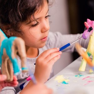 A close-up of a girl sitting next to another girl while they work on painting a unicorn figurine with different colors.