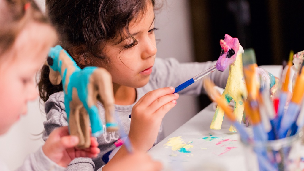 A close-up of a girl sitting next to another girl while they work on painting a unicorn figurine with different colors.