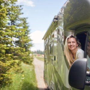 A woman is sticking her head out a window as she sits in the passenger’s seat of an RV. The RV is in a wooded area.