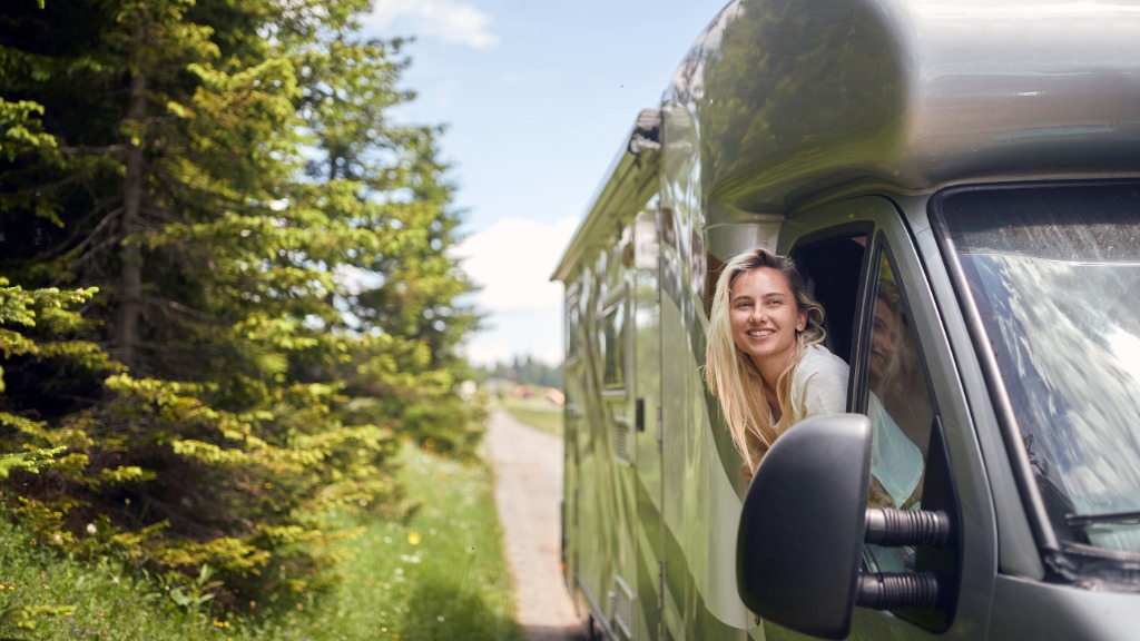 A woman is sticking her head out a window as she sits in the passenger’s seat of an RV. The RV is in a wooded area. A woman is sticking her head out a window as she sits in the passenger’s seat of an RV. The RV is in a wooded area.