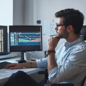 A businessman sits in his office, looking at various charts on his computer screens. He wears glasses and a blue shirt.