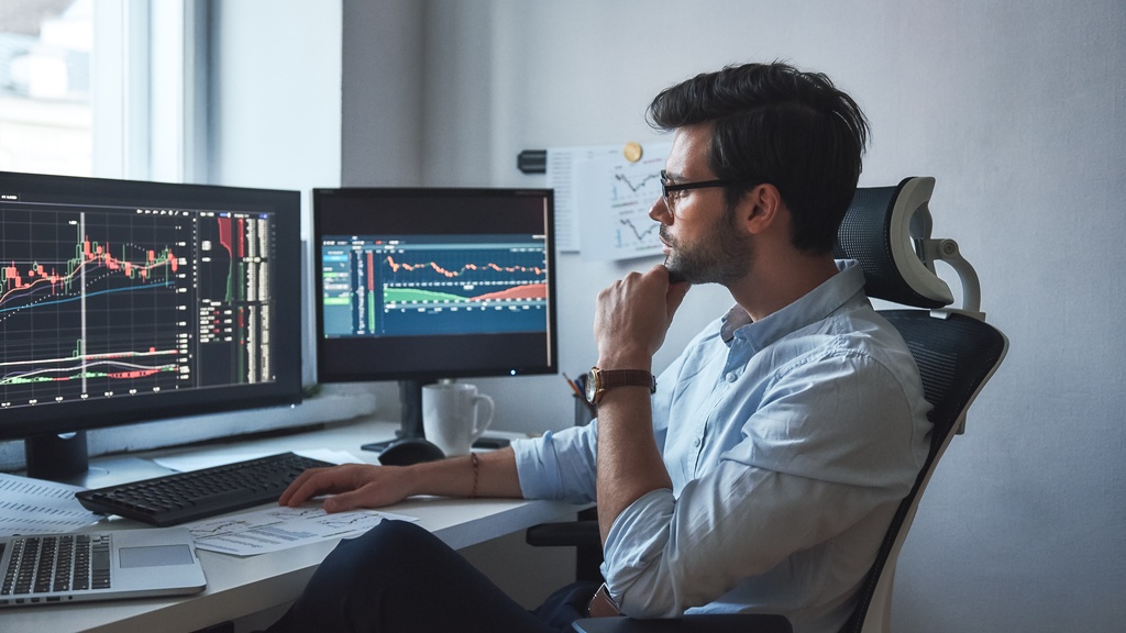 A businessman sits in his office, looking at various charts on his computer screens. He wears glasses and a blue shirt.